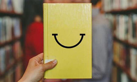Photographie d'une main qui tient un livre. La couverture est jaune et un sourire est dessiné.
